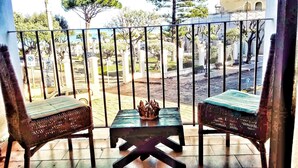 Un balcon avec deux fauteuils en osier et une petite table, donnant sur un cimetière où l’on aperçoit des pierres tombales blanches et des arbres.