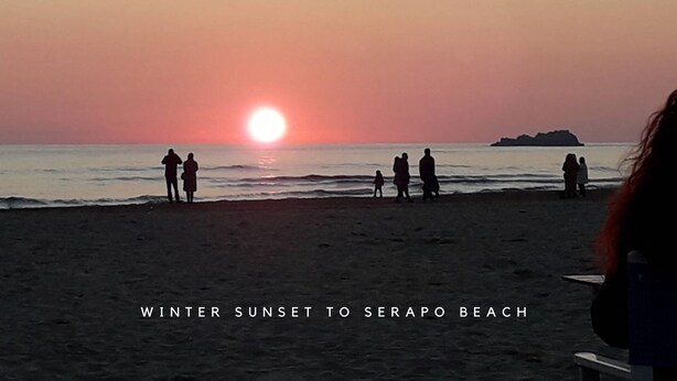 Un coucher de soleil d’hiver sur la plage de Serapio, avec des personnes debout sur le sable.