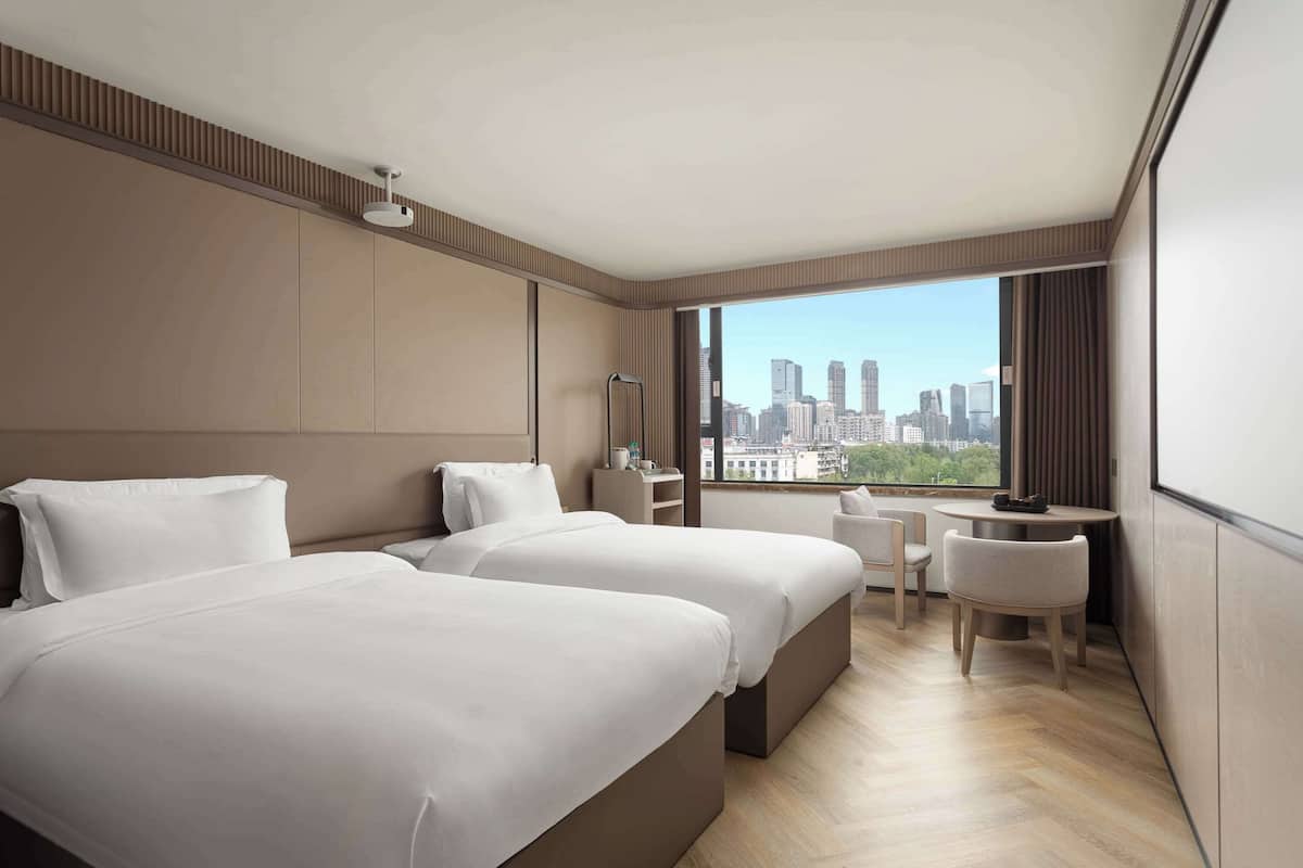 A modern hotel room with two beds, a desk, and a view of a city skyline.