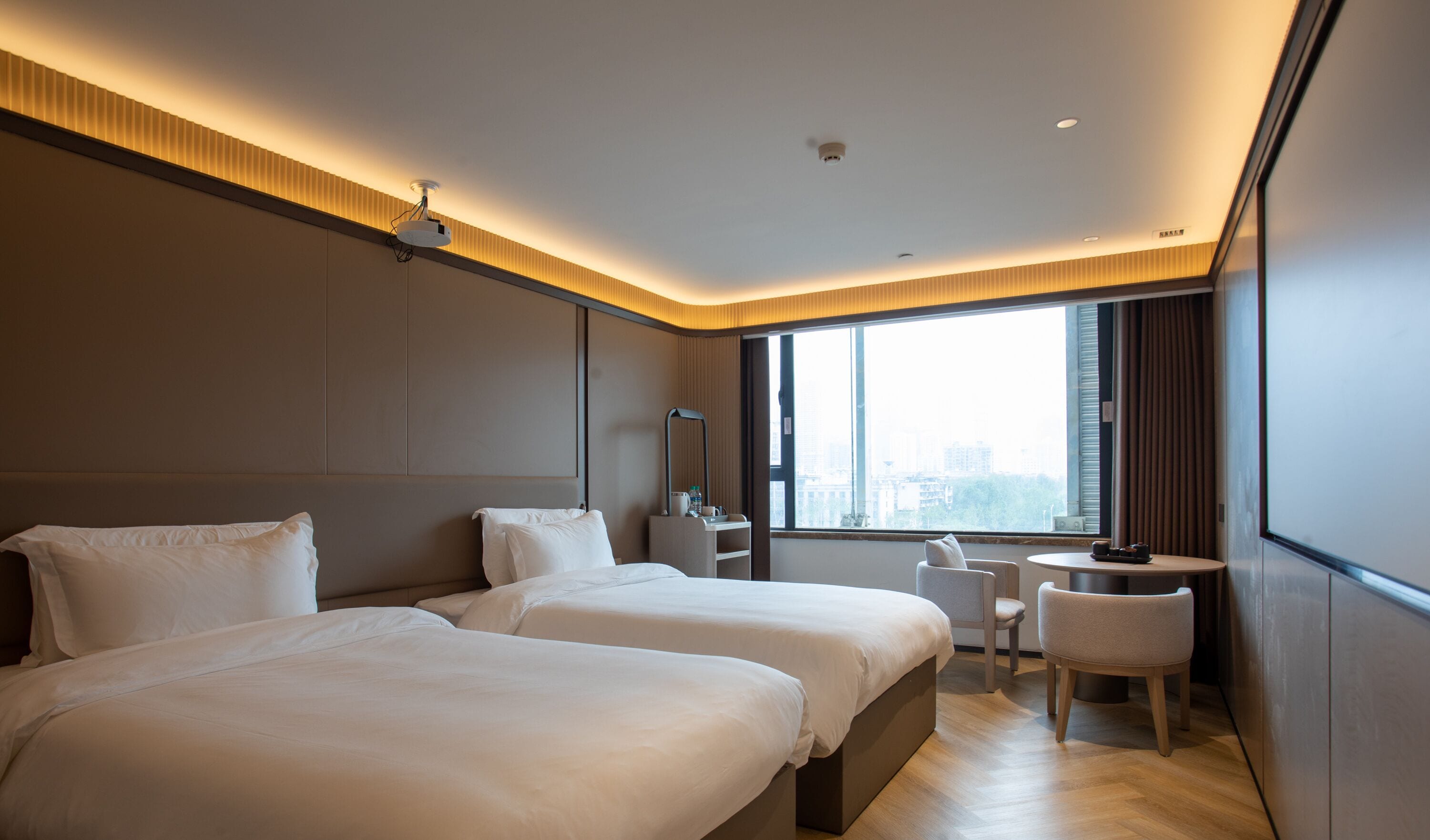 A modern hotel room with two beds, a desk, a chair, and a large window offering a city view.