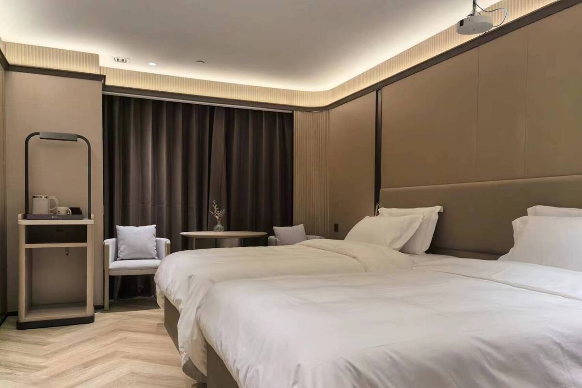 A modern hotel room with a large bed, a small table, a chair, and a desk with a lamp.