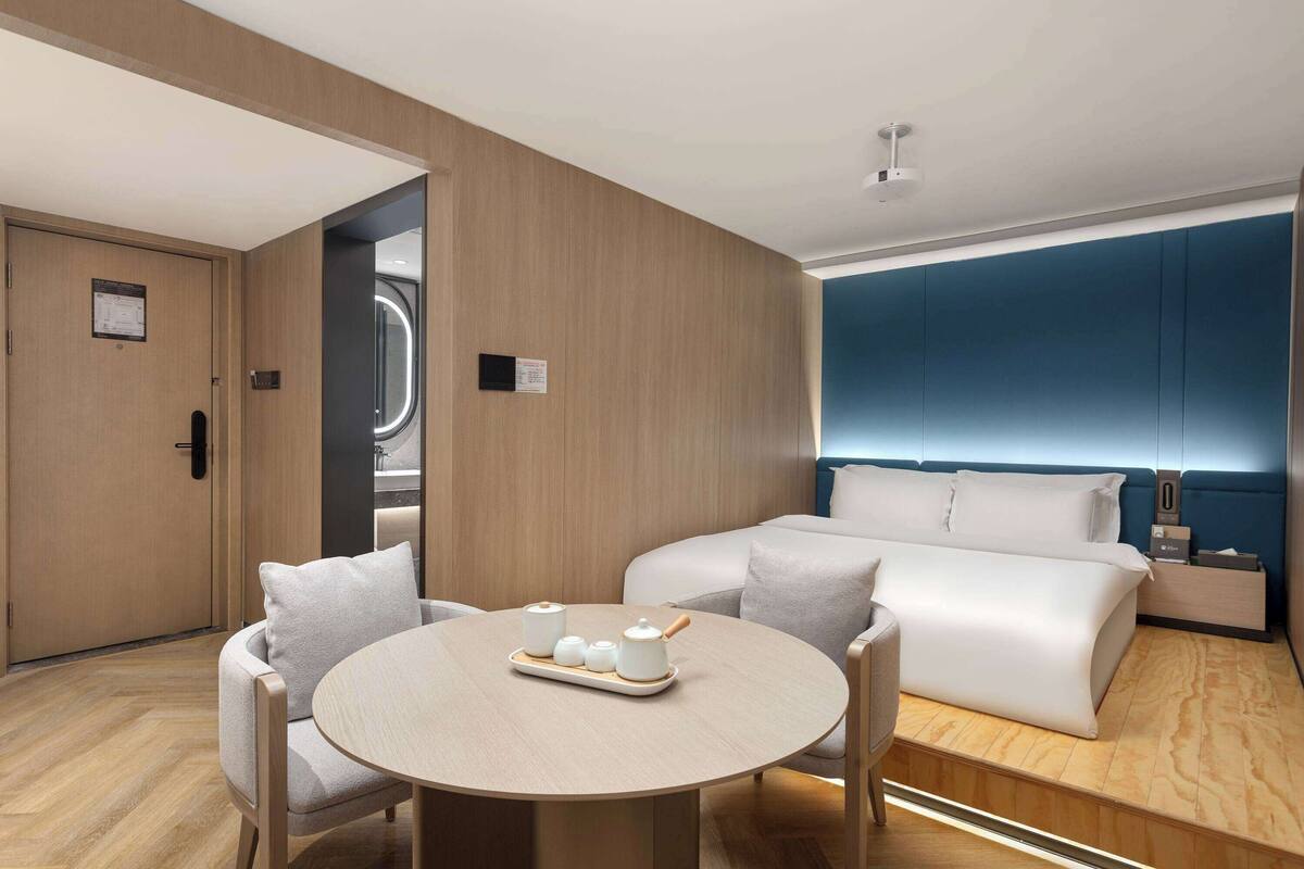 A modern hotel room with a wooden floor, a round dining table with a tray of tea set, a sofa, and a bed with a blue headboard.