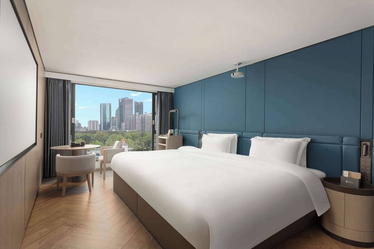 A modern hotel room with a large bed, a desk, and a chair, a view of the city, and a blue accent wall.