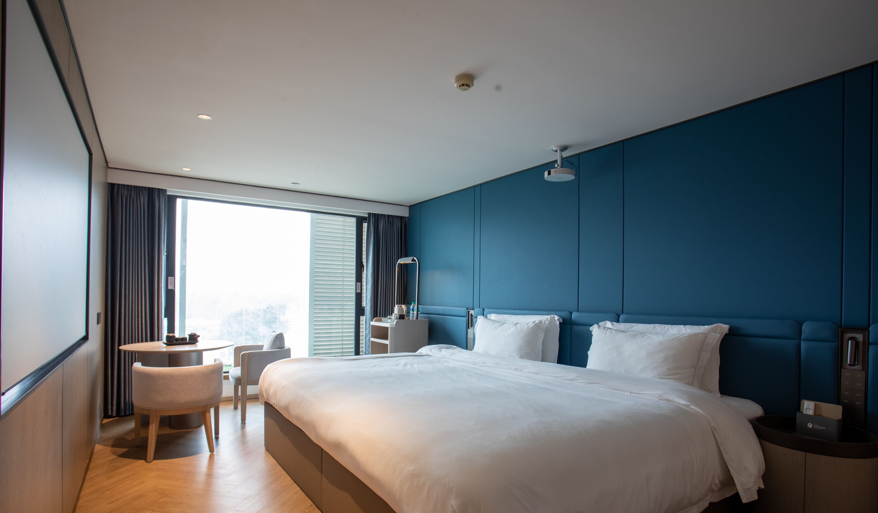 Ett modernt hotellrum med en stor säng, ett litet skrivbord, en stol och ett fönster med gardiner.