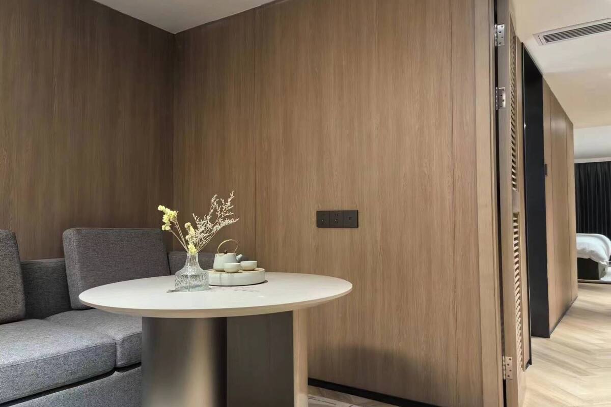 A modern hotel room with a grey sofa, a round table with a vase of flowers, and wooden paneling on the walls.
