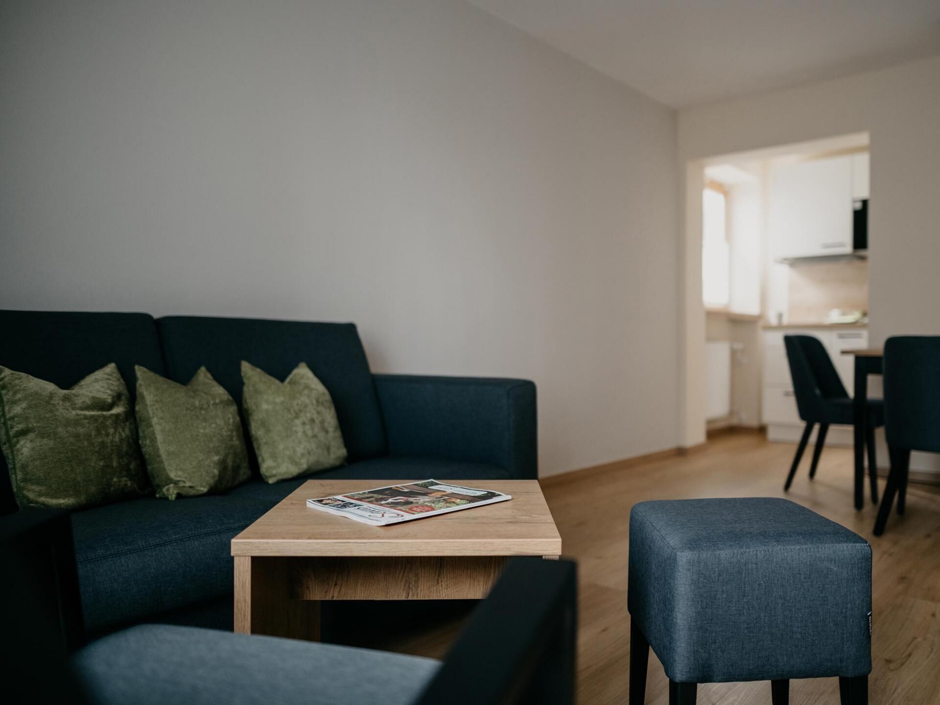 Ein modernes Wohnzimmer mit einem dunkelblauen Sofa, einem Holztisch und passendem Hocker. Eine Zeitschrift liegt auf dem Tisch.