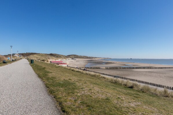 Une route côtière menant à une plage, bordée d’une clôture en bois et donnant sur un bâtiment au toit rouge.