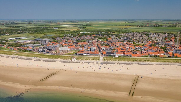 Vue aérienne d’une ville côtière avec une plage, des b âtiments aux toits rouges et, au loin, de vastes champs.