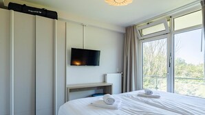 A hotel room with a bed, a flat-screen TV mounted on the wall, a window with curtains, and a view of trees outside.