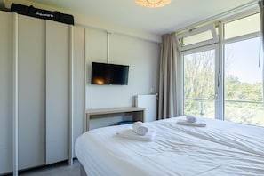 A hotel room with a bed, a flat-screen TV mounted on the wall, a window with curtains, and a view of trees outside.