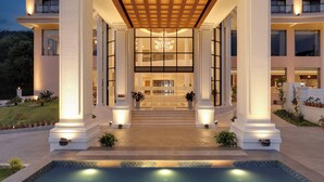 Une entrée d’hôtel moderne avec une piscine, de grandes colonnes et un imposant escalier menant au hall d’entrée.