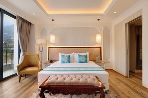Relax in this serene hotel room with a large bed, wooden bench, and armchair, offering a stunning mountain view.