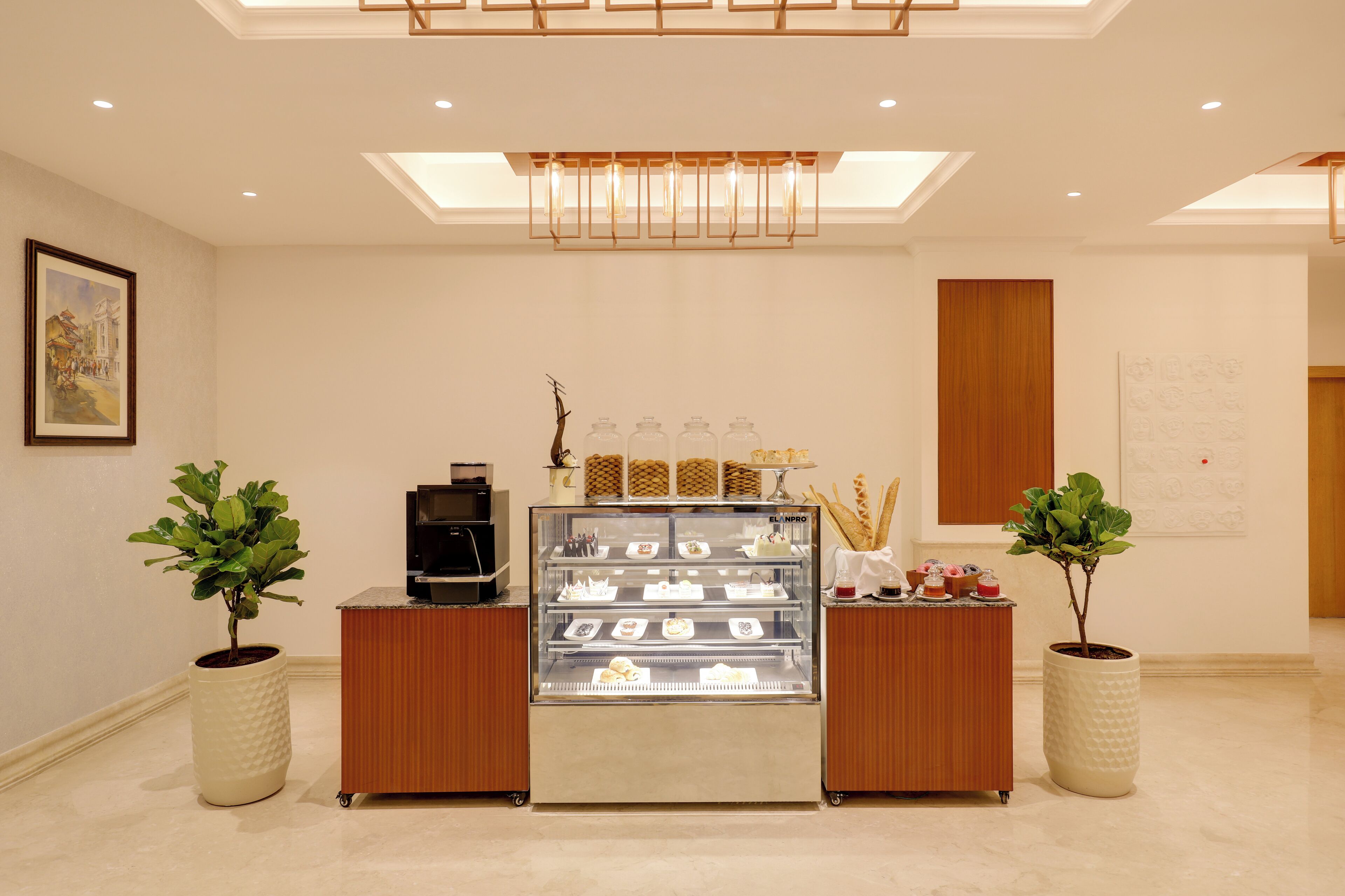 A modern hotel lobby with a bakery display, potted plants, and a framed artwork on the wall.