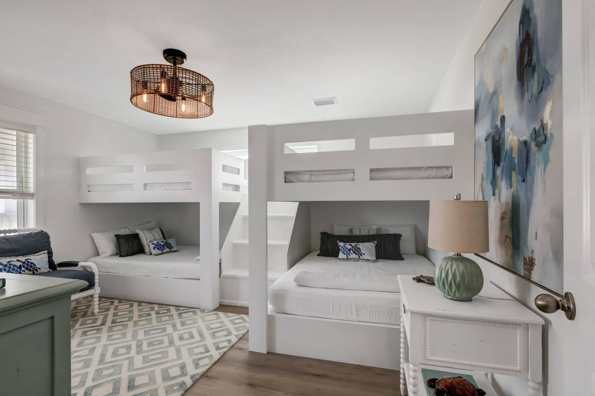 A modern bedroom with two bunk beds, a patterned rug, a side table with a lamp, and a painting on the wall.