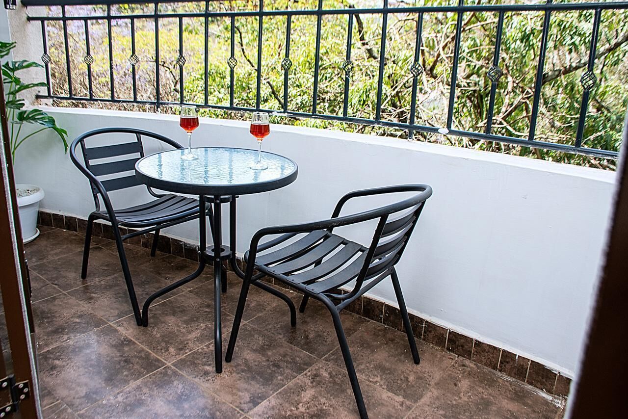 Um pequeno pátio ao ar livre com uma mesa redonda e duas cadeiras, um vaso de planta e vista para o verde além de uma grade de metal.