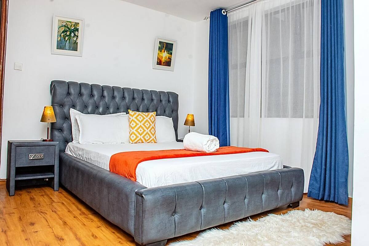 Ein Schlafzimmer mit einer gepolsterten Kopfteile, einem Bett mit weißer und orangefarbener Bettwäsche, zwei Nachttischlampen, einem kleinen Nachttisch und einem flauschigen weißen Teppich.