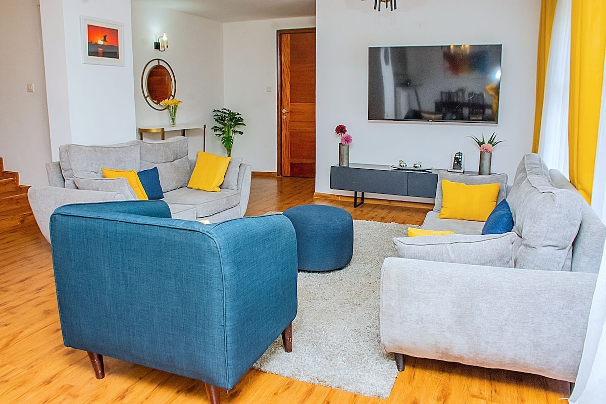 Ein modernes Wohnzimmer mit einem grauen Sofa, einem blauen Sessel und einem grauen Sessel mit gelben Kissen. Ein weißer Teppich, Holzboden und ein Fernseher an der Wand sind ebenfalls vorhanden.