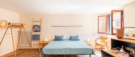 A bedroom with a bed, a wooden chair, a small table, a radiator, and a shelf with books and a lamp.