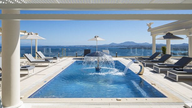 Piscina infinita no terraço com fonte de água, cercada por espreguiçadeiras e guarda-sóis, com vista para uma paisagem montanhosa.