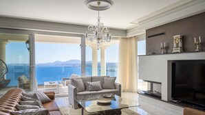 Uma sala de estar ampla com uma televisão grande, assentos confortáveis, uma mesa de centro de vidro e vista para o mar.