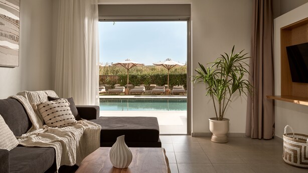 A modern living room with a sofa, a wooden coffee table, a potted plant, and a view of a pool and outdoor seating area.