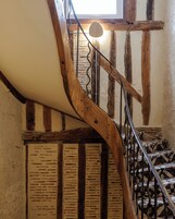 Una escalera espiral de madera con barandilla de metal, que conduce a una habitación con paredes de ladrillo visto y un ventanal.