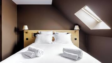 A hotel room with a bed, two bedside lamps, a skylight, and two rolled towels on the bed.