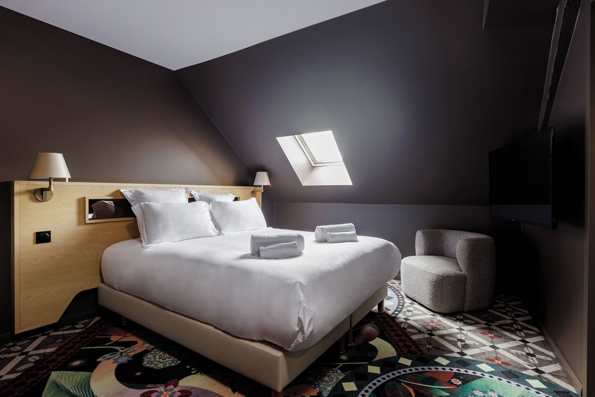 Una habitación de hotel moderna con una cama grande, un sillón pequeño, un tragaluz y un televisor de pantalla plana.