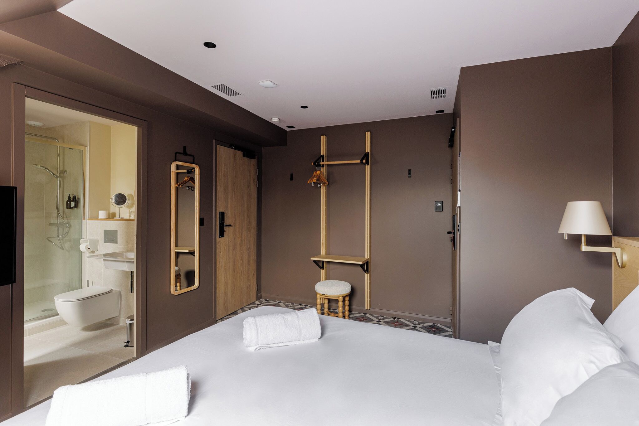 Une chambre d’hôtel moderne avec un grand lit, un tabouret en bois, un miroir et un espace douche visible à travers une porte ouverte.