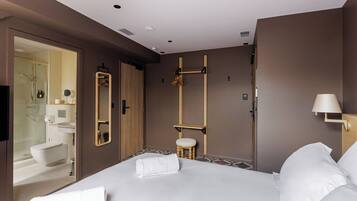 Une chambre d’hôtel moderne avec un grand lit, un tabouret en bois, un miroir et un espace douche visible à travers une porte ouverte.