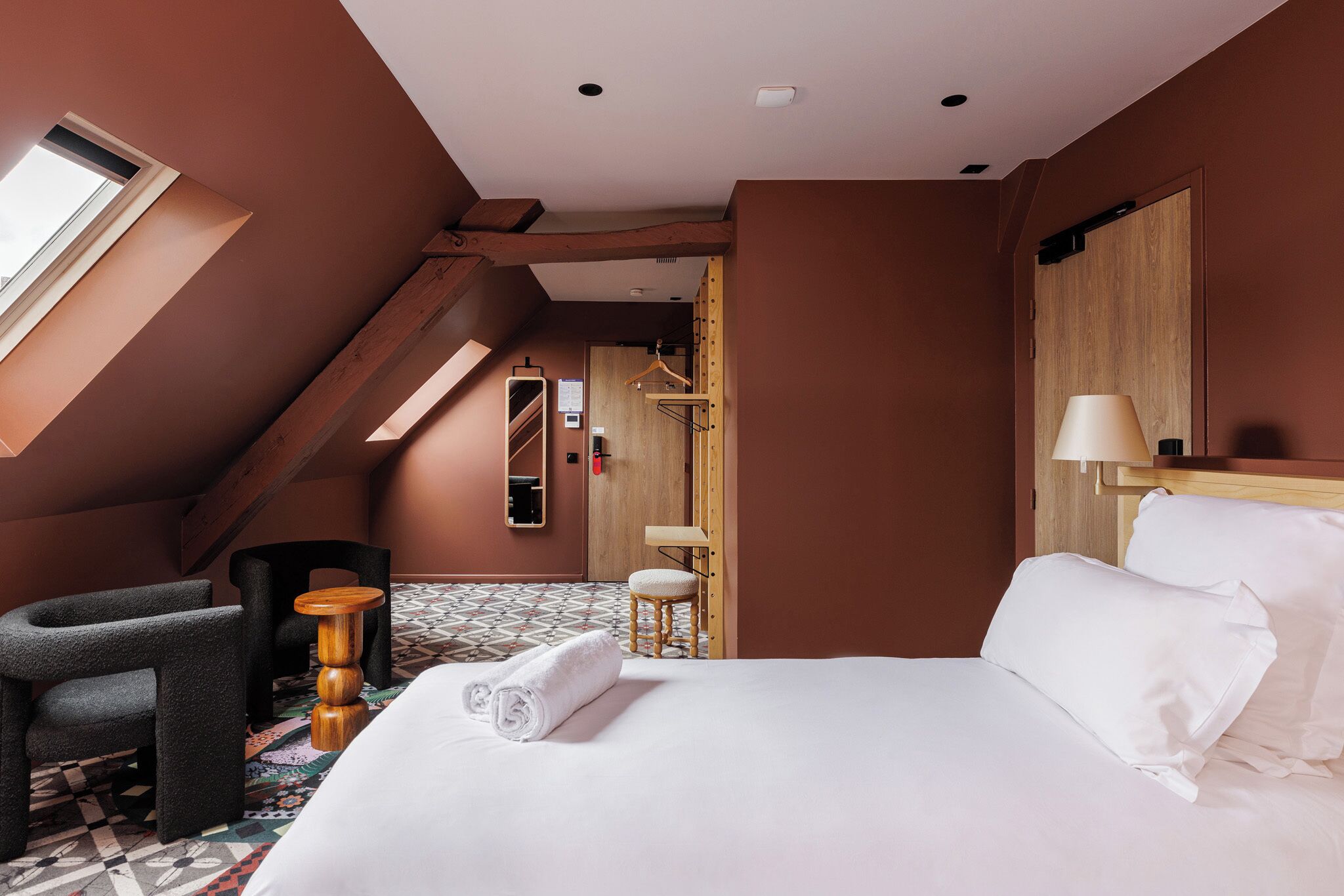 Una habitación de hotel con cama, una silla, un taburete pequeño, una lámpara colgante y una ventana.