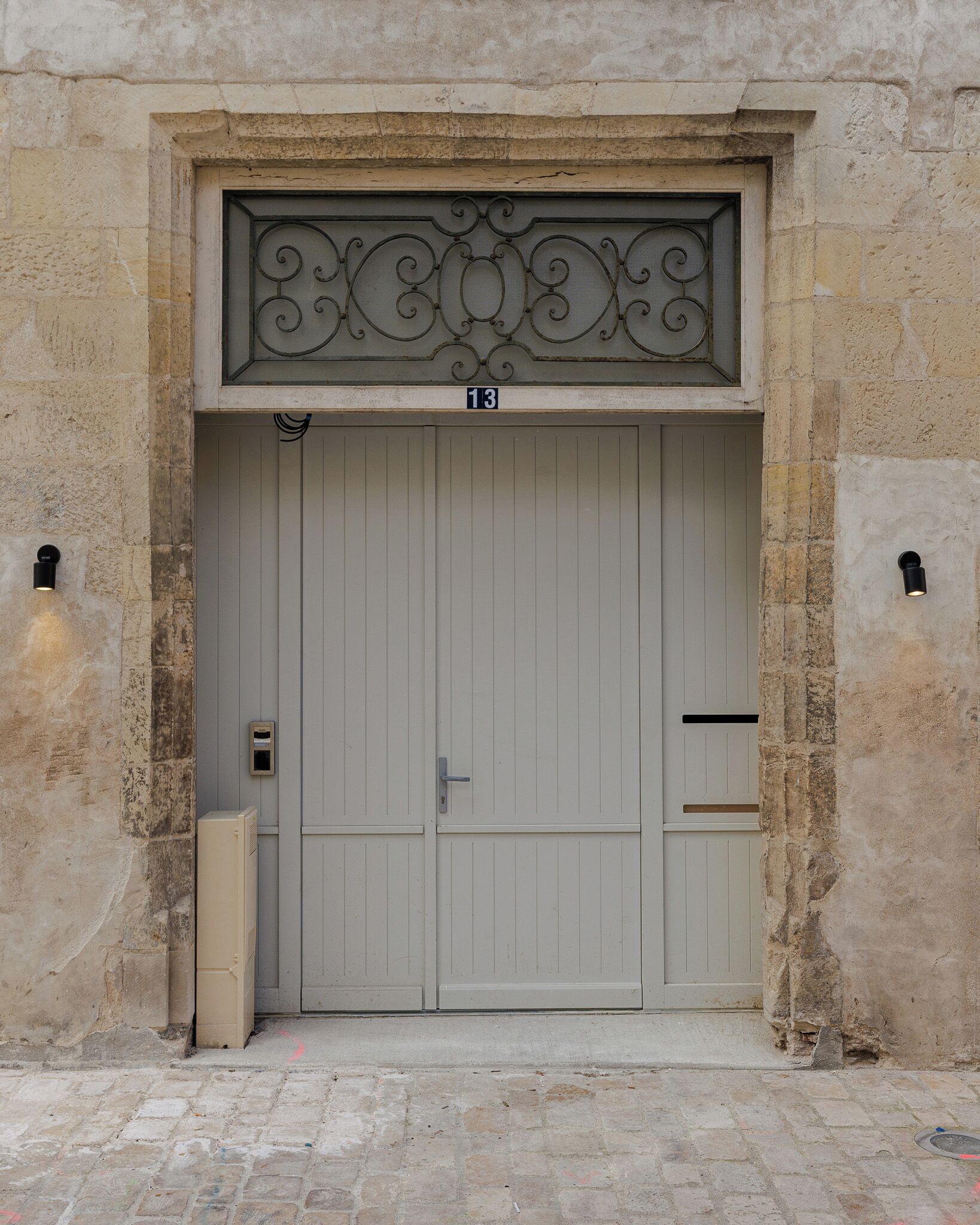 Une grande porte à deux battants, surmontée d’une ornementation métallique décorative, le tout intégré dans un mur de pierre.