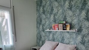 Um quarto com uma cama, um ventilador de teto, uma janela com persianas, uma parede com papel de parede estampado com folhas e uma estante com livros e um par de óculos.