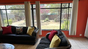 A living room with two black sofas, one with red and yellow cushions, and a round coffee table. Large windows with curtains offer a view of a garden. - The White House  (Phalaborwa)