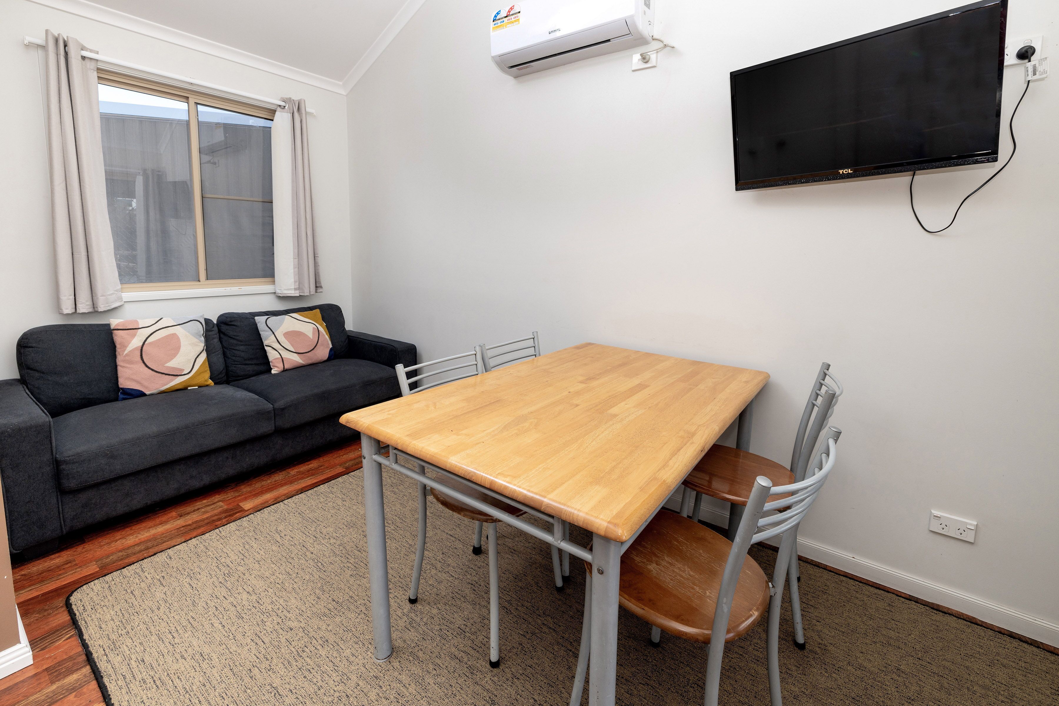A modern living room with a grey sofa, a wooden dining table, and a TV mounted on the wall.