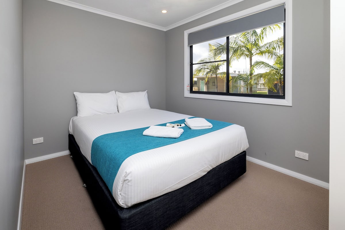 Een net opgemaakt bed met een blauw-witte spreideken, een raam met uitzicht op palmbomen en een grijze muur.