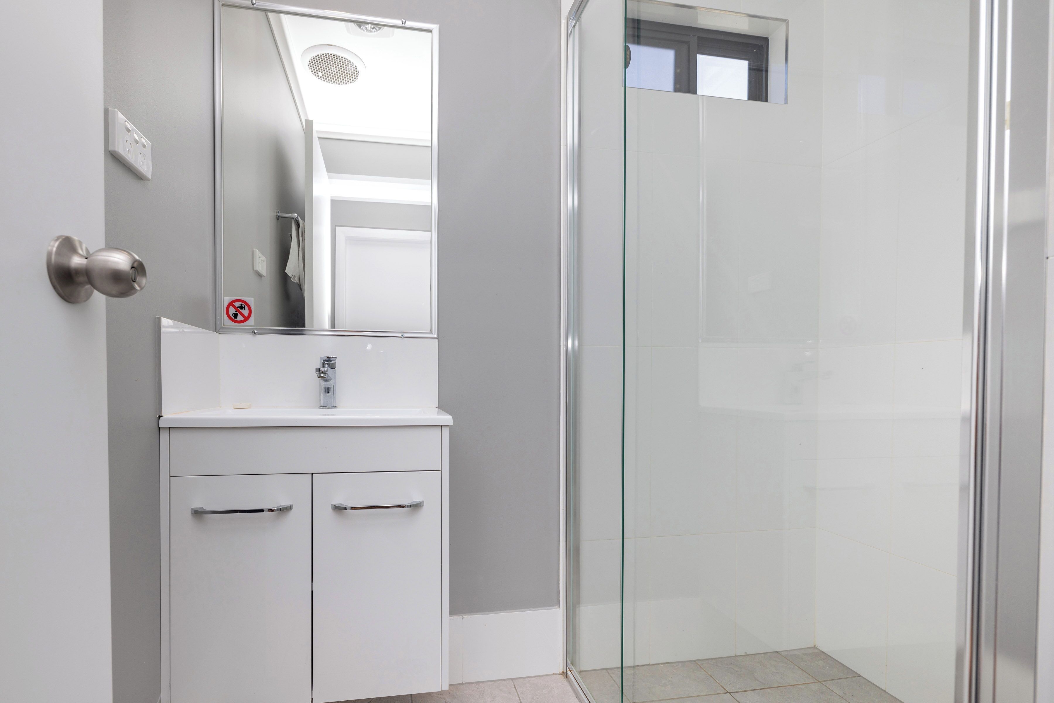 Une salle de bain avec une douche à parois de verre, un lavabo blanc avec un miroir et une porte sur laquelle est apposé un panneau « interdiction de fumer ».