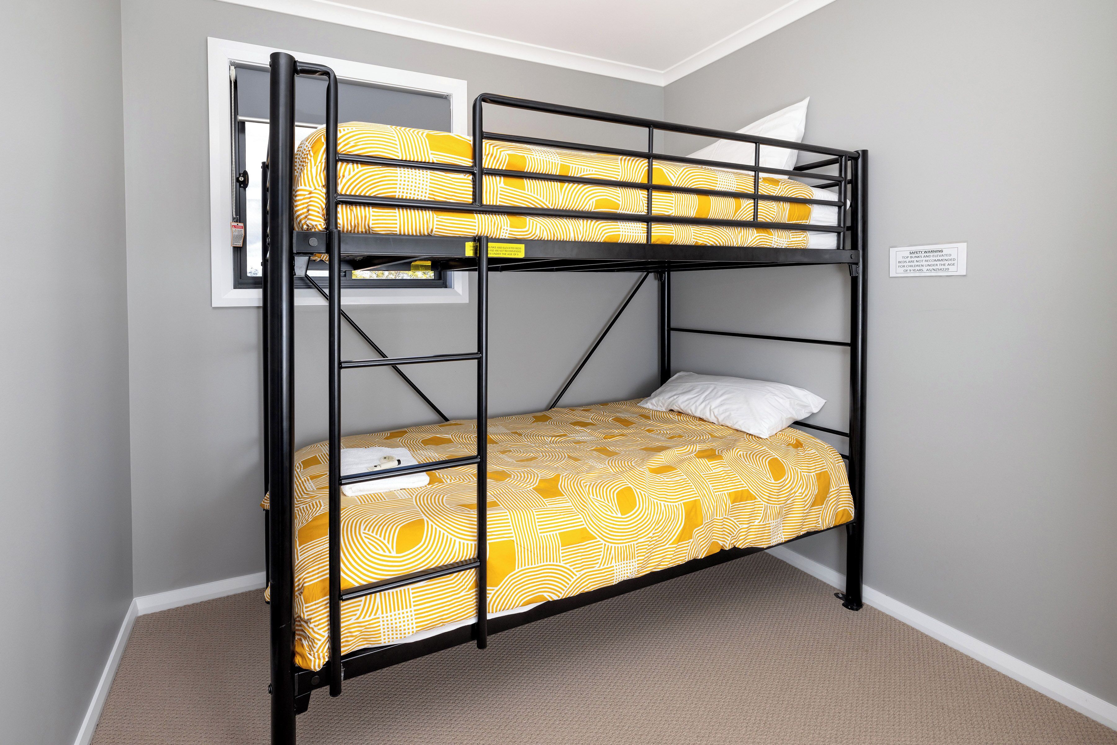 Un lit superposé avec une couvre-lit à motifs jaunes et des oreillers blancs, une fenêtre donnant sur un paysage agréable et un panneau d’avertissement de sécurité fixé au mur.