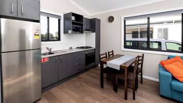 A modern kitchen with grey cabinets, a stainless steel refrigerator, a microwave, and a gas stove. There is a dining table with chairs and a blue couch with an orange throw pillow. A window provides a view of a parked car outside.
