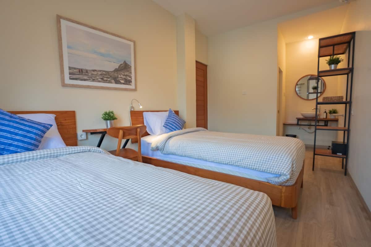 A bedroom with two beds, a bedside table with a lamp, a framed picture on the wall, and a small shelf with a plant.