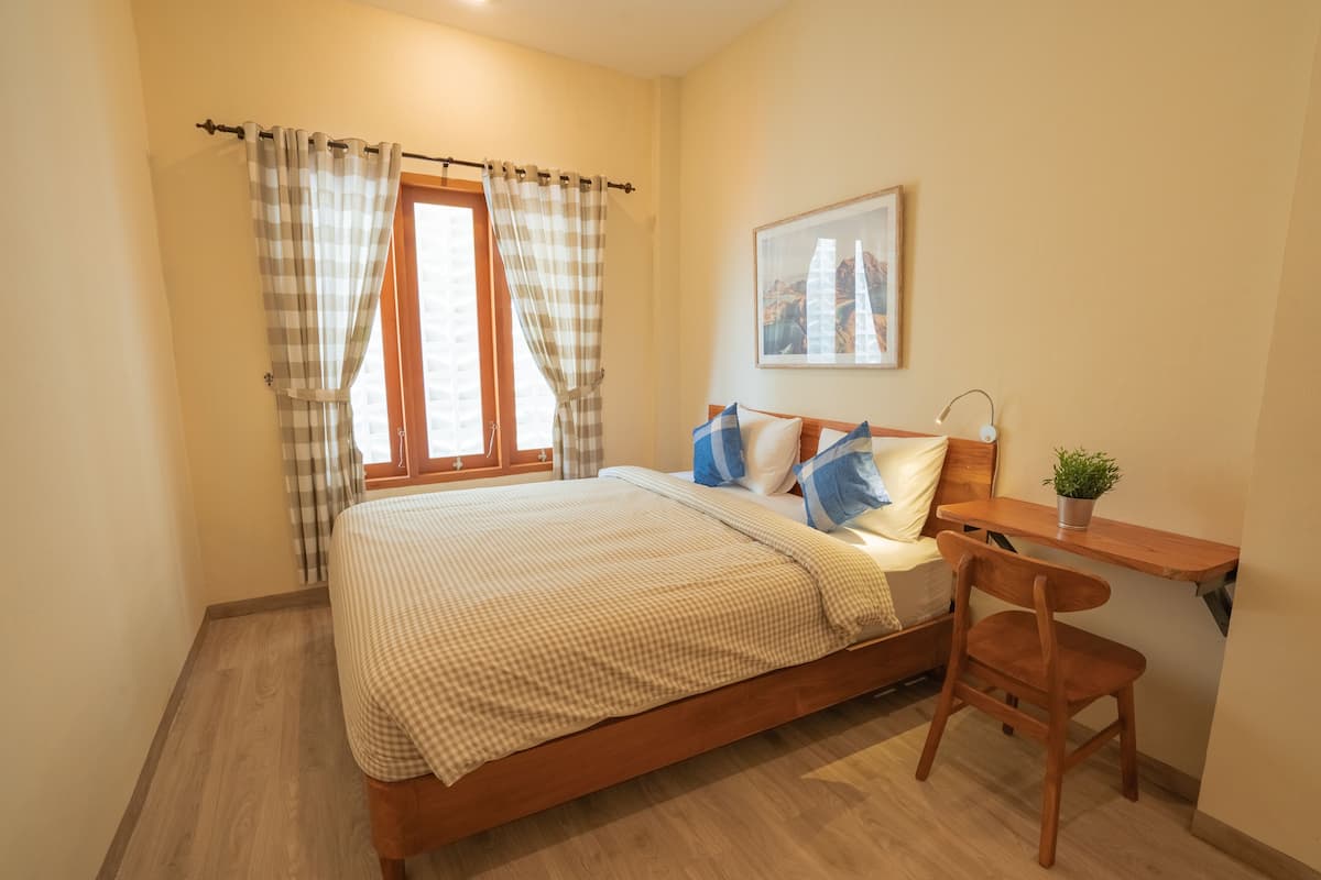 A bedroom with a bed, a desk, a chair, a window with curtains, and a framed picture on the wall.