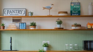 A kitchen with a green tiled backsplash, a wooden countertop, and various kitchen appliances. There are potted plants on the countertop and a sign that reads "PANTRY OPEN 24 HOURS".