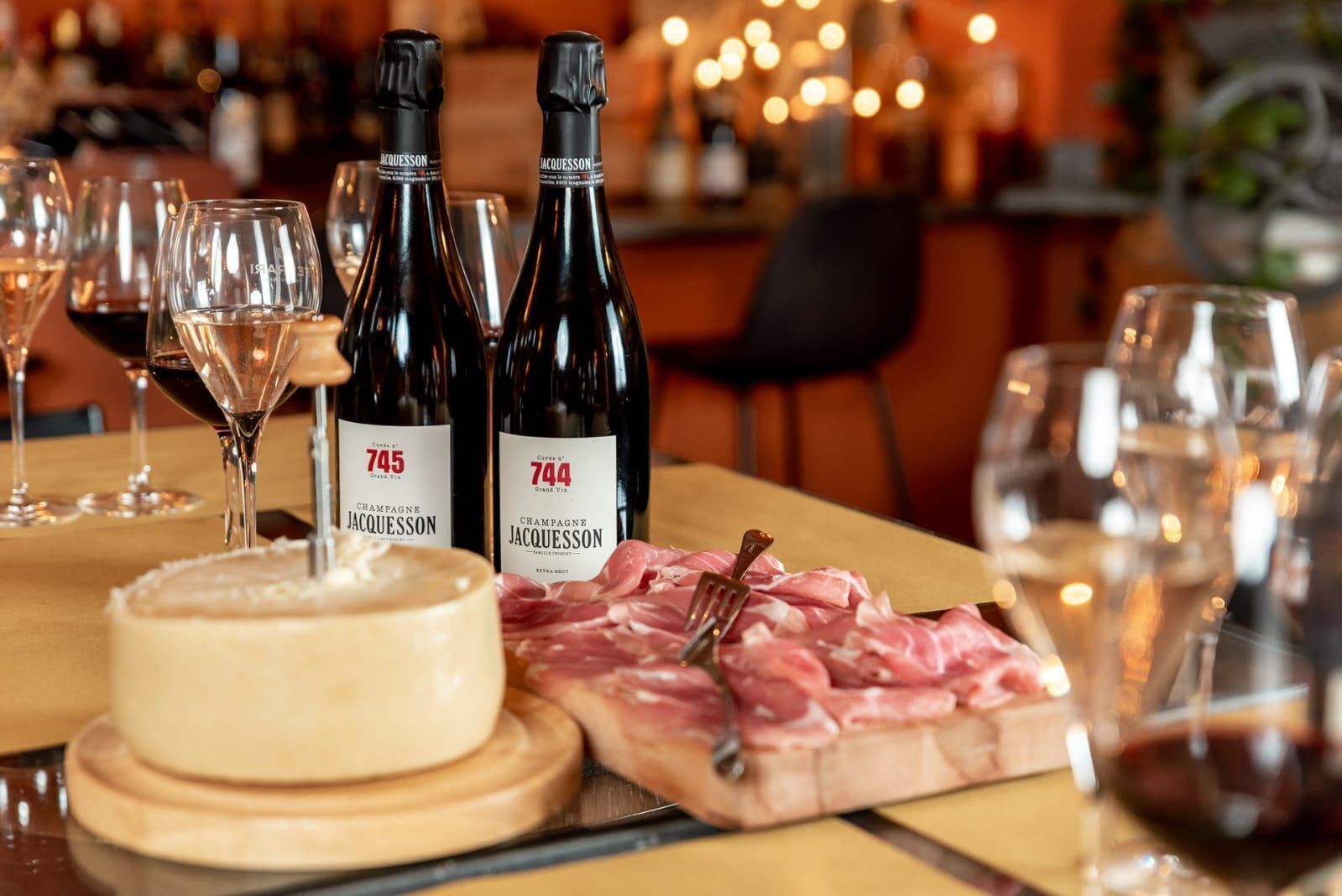 A dining table with two bottles of Jacquesson Champagne, a round cheese wheel, and a board of sliced meats.