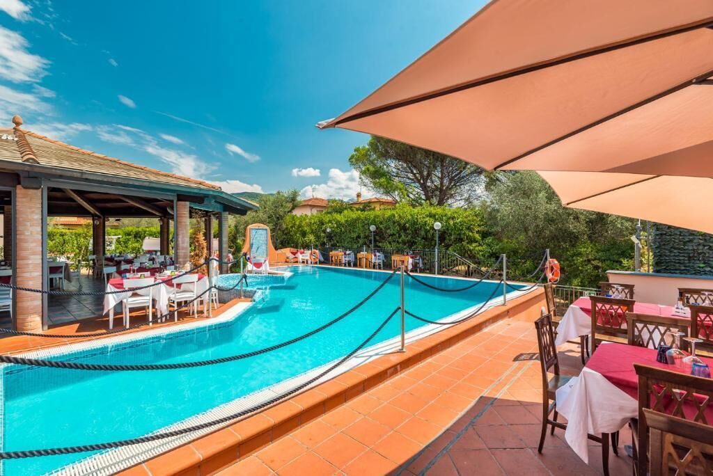 A pool area with a covered seating area and tables, surrounded by greenery and a clear sky.