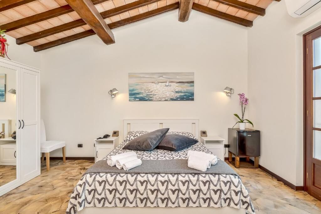 A bedroom with a patterned bedspread, a painting of sailboats, bedside lamps, a small plant, and a wooden ceiling.