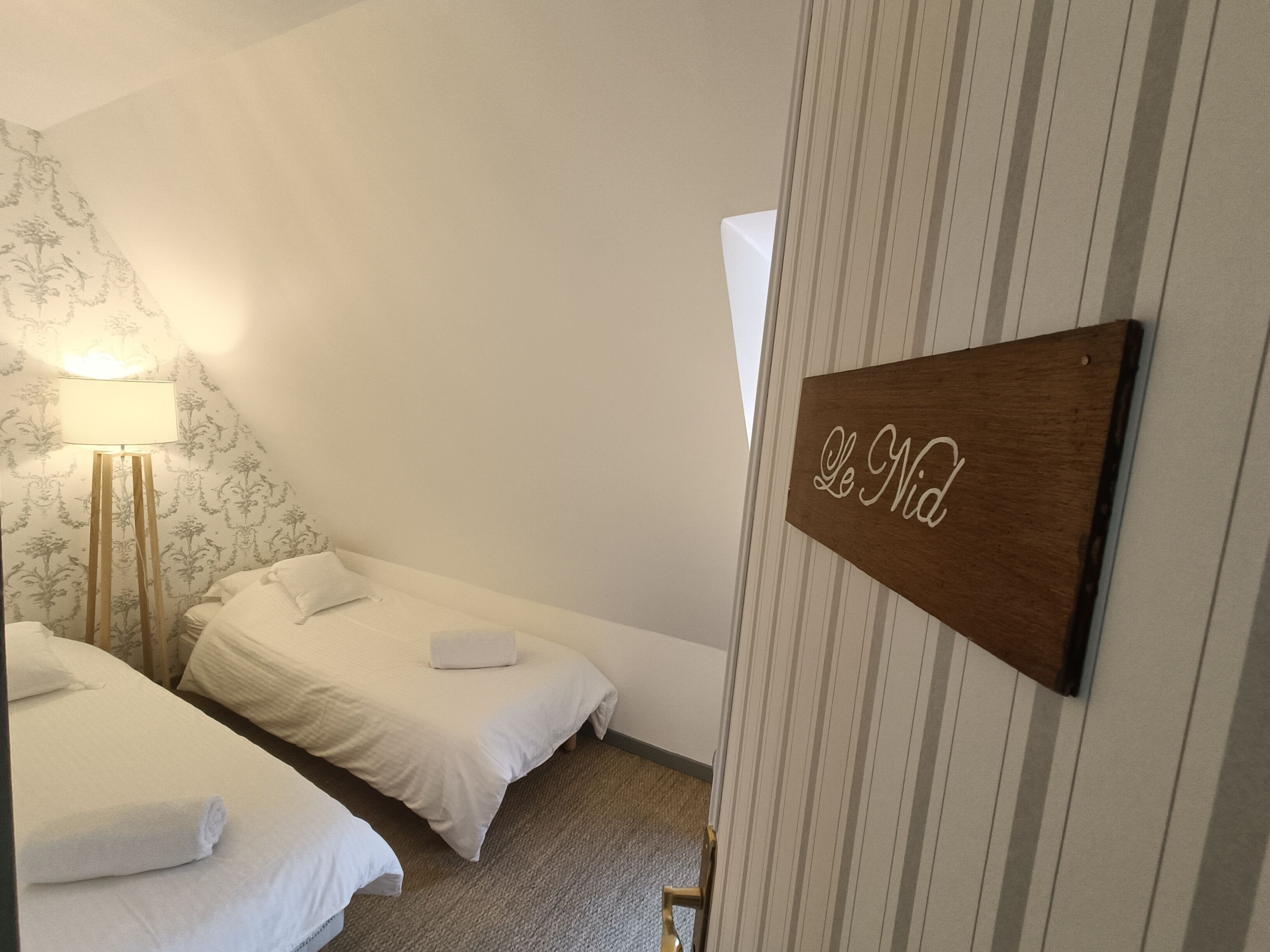 Een hotelkamer met twee bedden, een bedlampje, een houten bordje met 'Le Nid' en een gestreepte muur.