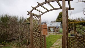 A wooden arbor gate leading to a garden area with a greenhouse and a brick structure. - Red Brick Former Railway Workers' Cottage (Brora)