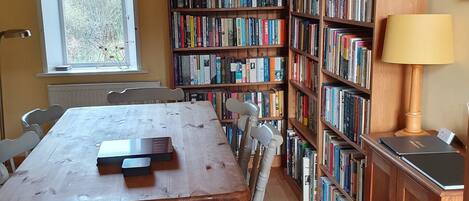 Um quarto com uma mesa e cadeiras de madeira, uma estante repleta de livros, uma janela e um abajur.