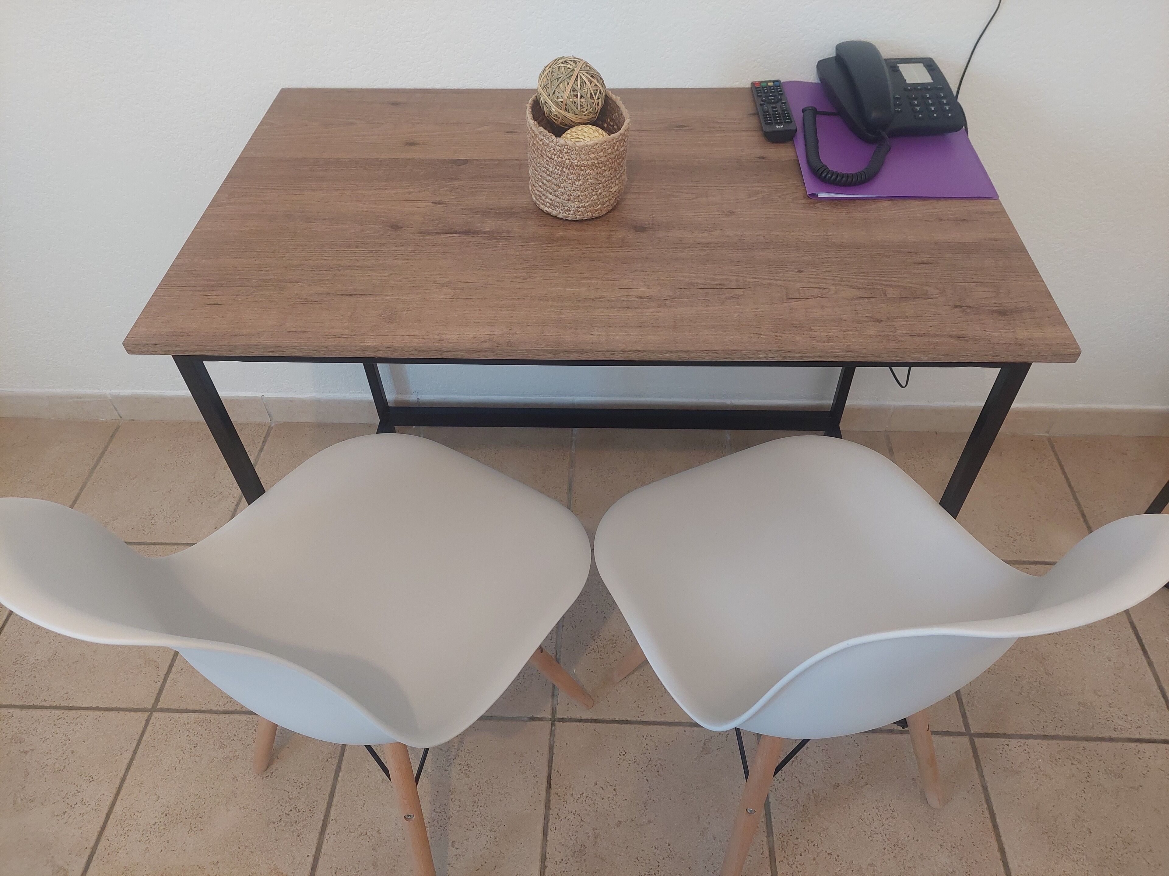 A wooden table with a woven basket and a telephone. Two white chairs are positioned in front of the table.