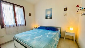 Un dormitorio con cama, dos mesitas de noche, una ventana con cortinas, un cuadro en la pared y una mesita de noche con lámpara.